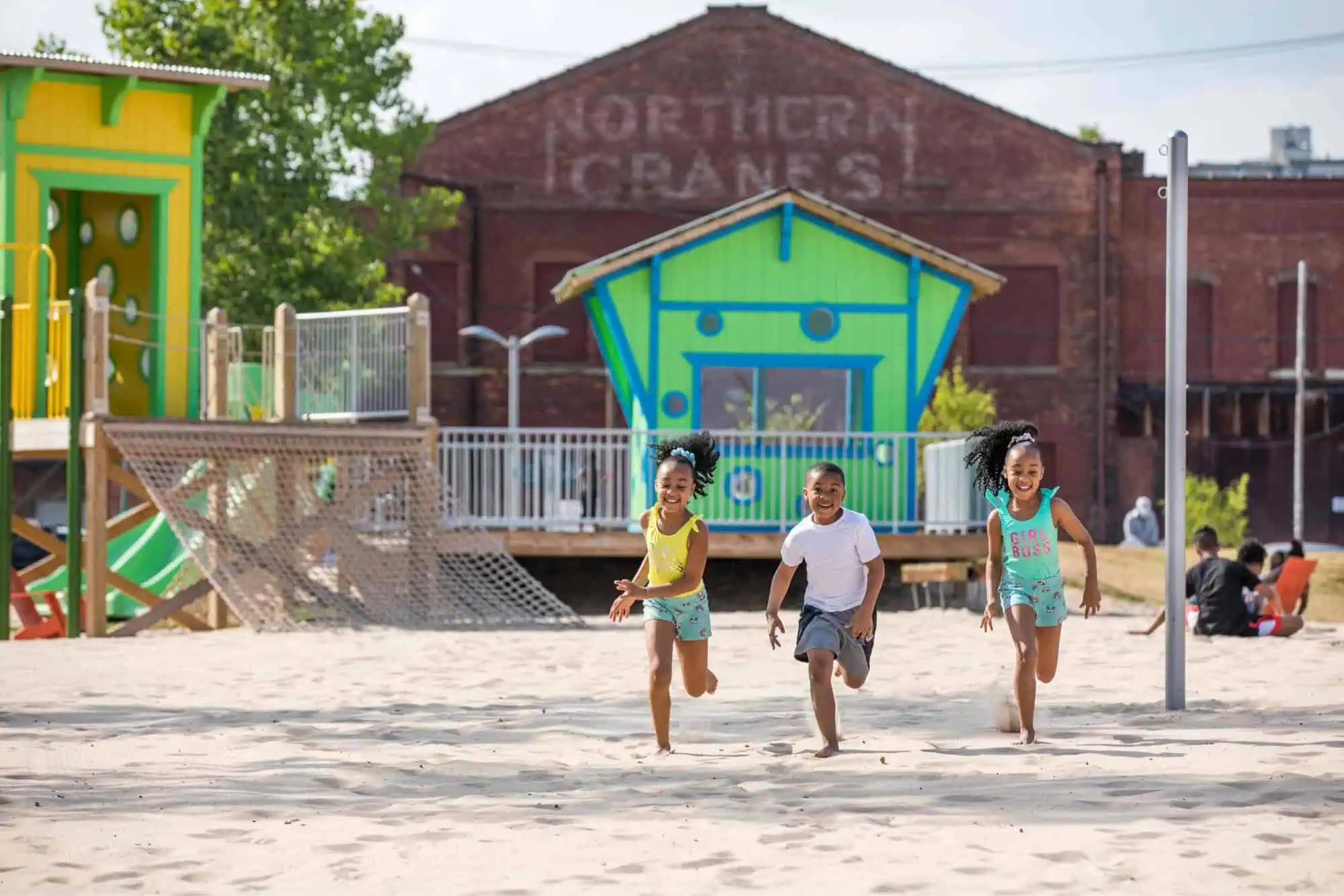 Three children running in the sand with a playground in the background