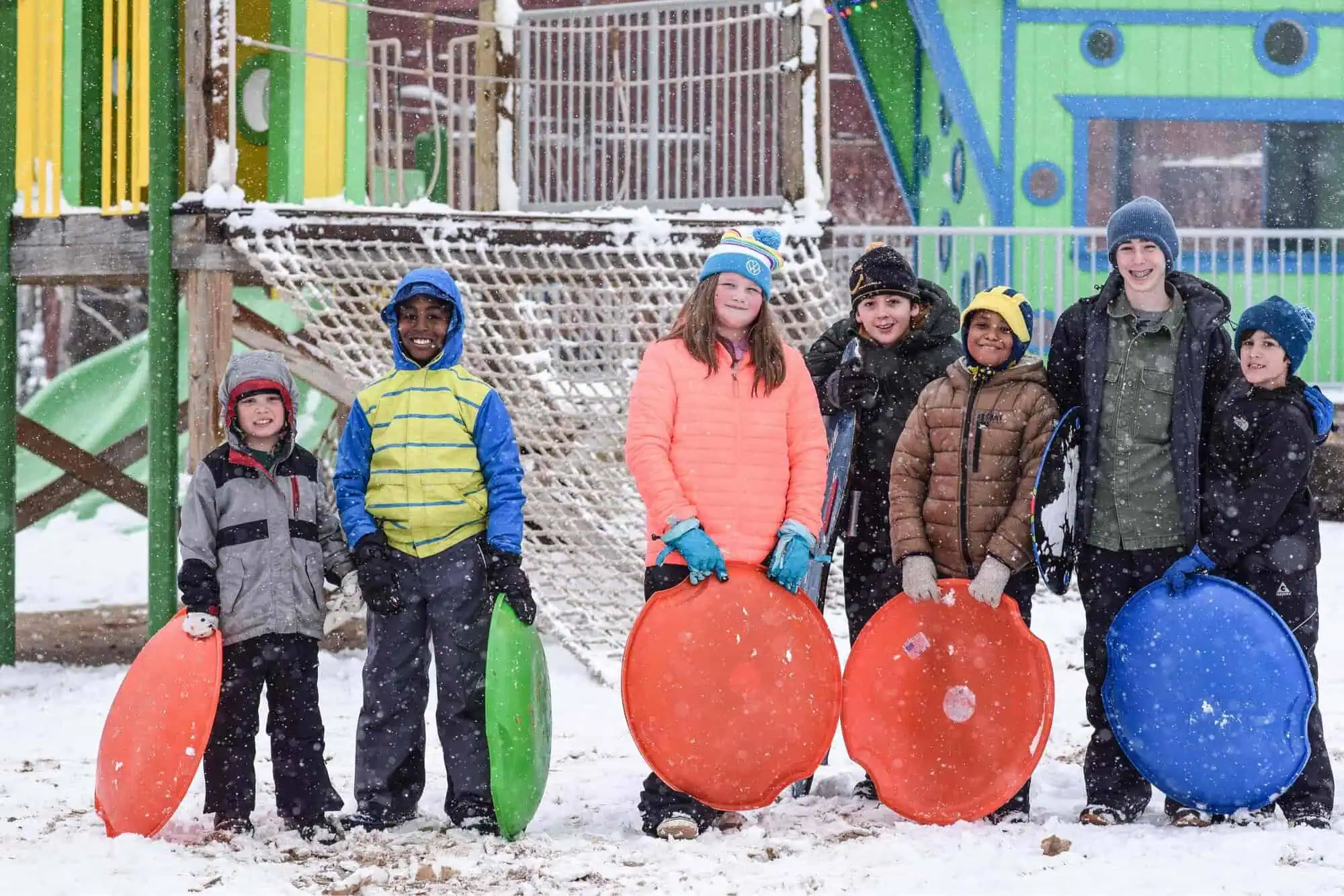 A group of kids in winter coats holding saucer sleds, with snow falling and snow on the ground