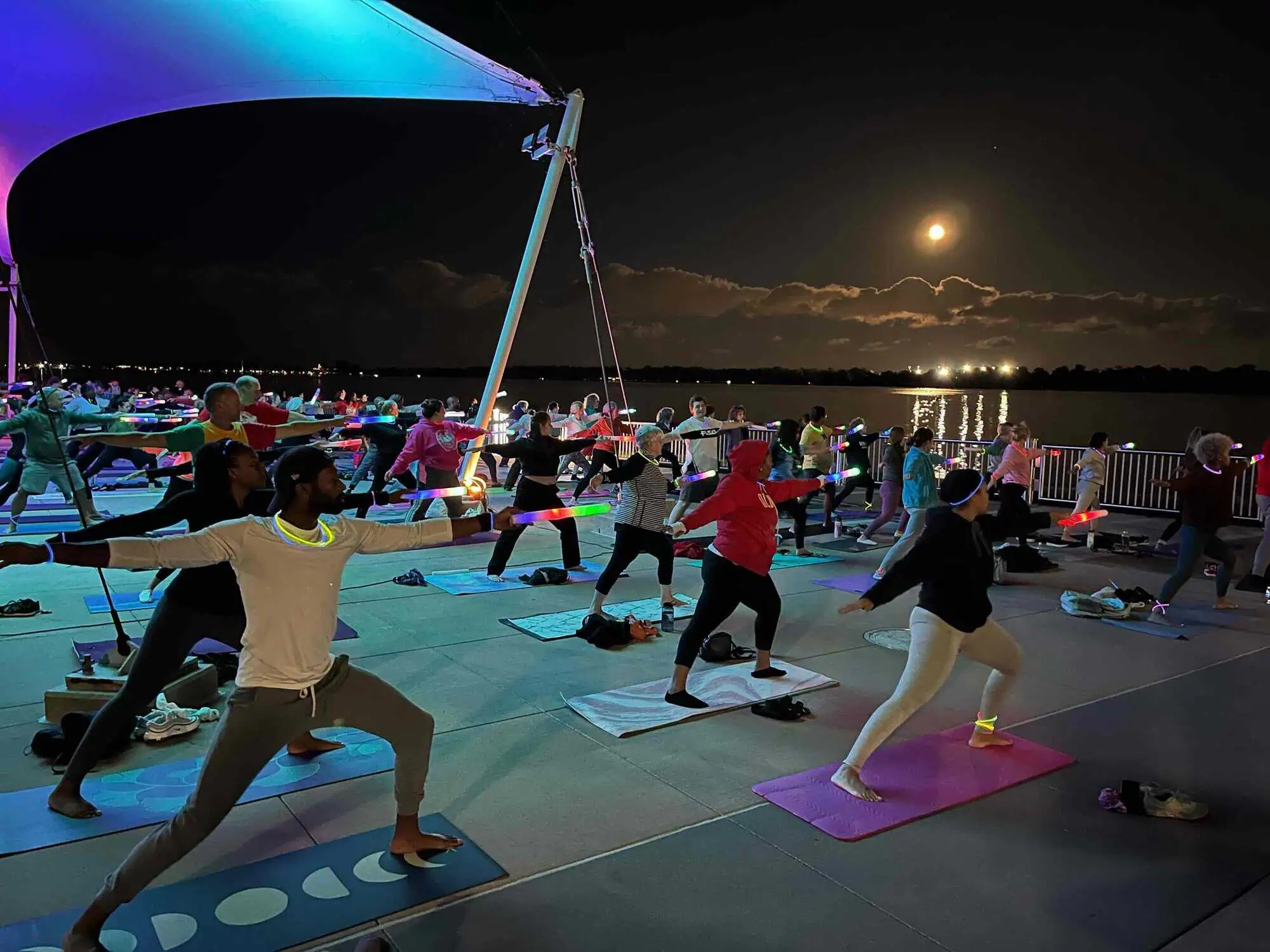 A large group of people at night practicing tai chi near the shore line. They are wearing and holding glowsticks.